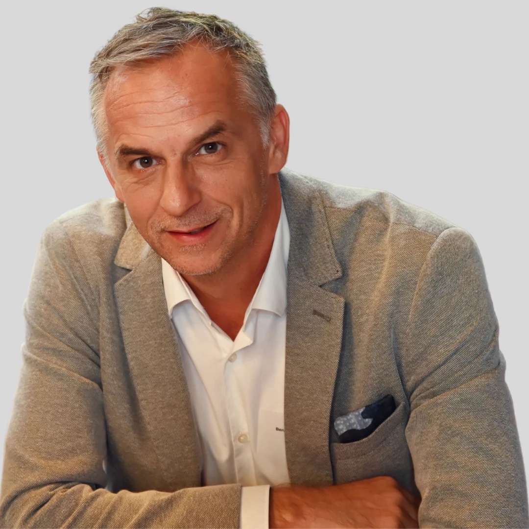 Portrait of a smiling middle-aged man with short gray hair, wearing a beige blazer and open white shirt, leaning forward with arms crossed.